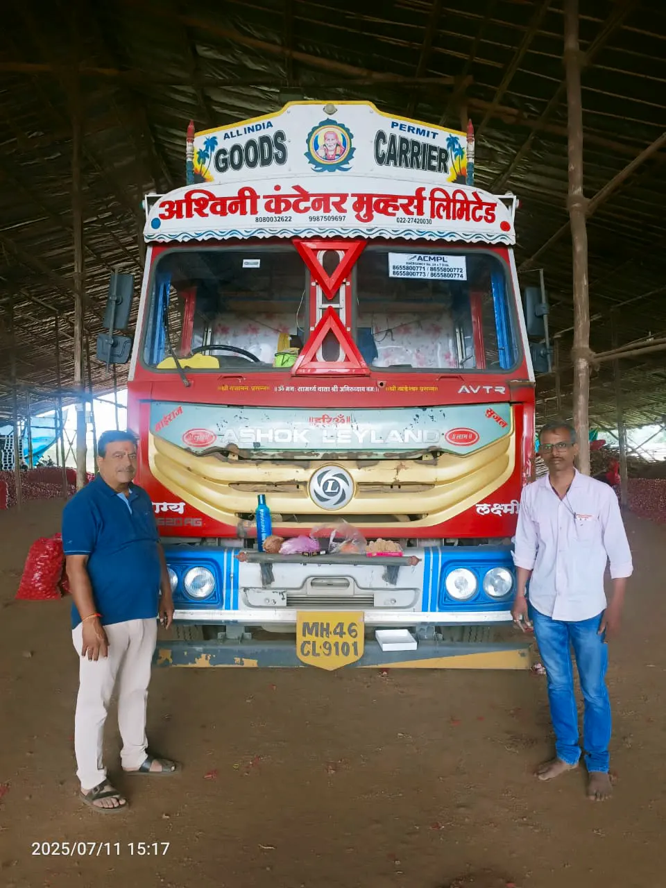 Two team members standing in front of a red Ashok Leyland truck with company name board
